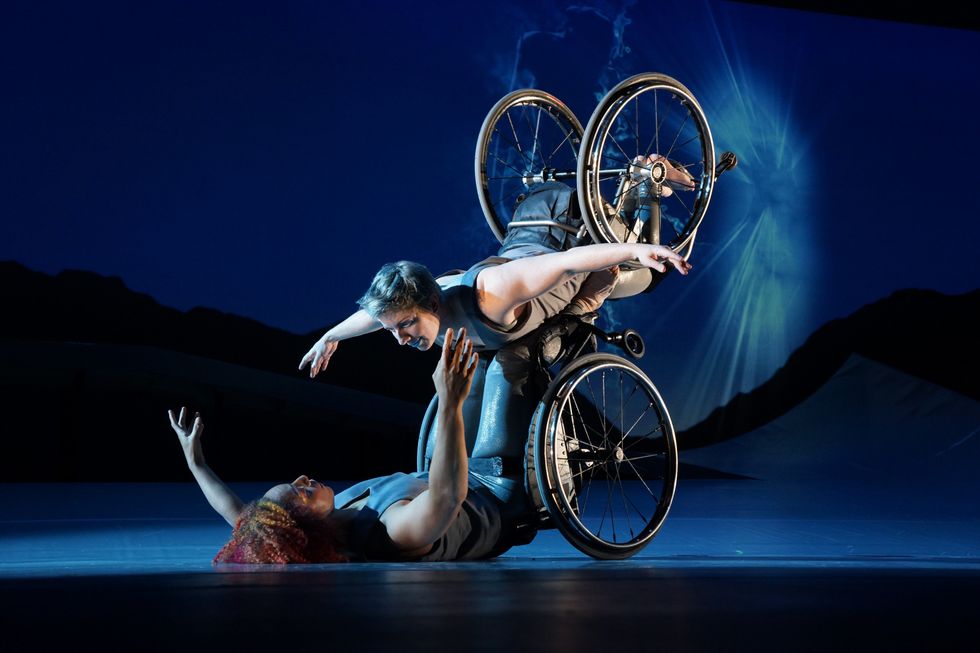 Laurel Lawson as Venus is flying in the air with arms spread wide, wheels spinning, and supported by Alice Sheppard as Andromeda who is lifting from the ground below. They are making eye contact and smiling. Photo by Jay Newman/BRITT Festival.