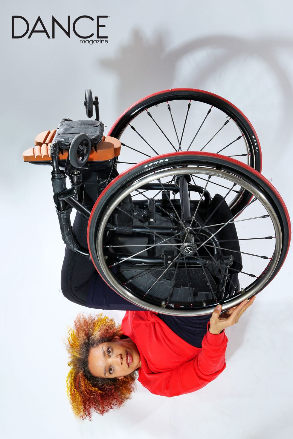 Alice Sheppard is upside down with wheels reaching to the sky. She looks directly at you. You can see the underside of her wheelchair, as though she is presenting it to you. Photo by Jayme Thornton.