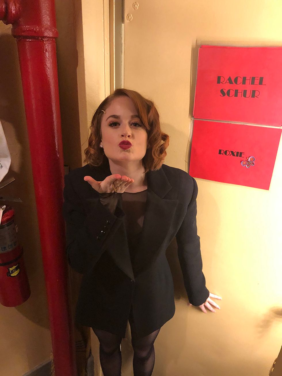 Schur standing in front of her dressing room door, which has a sign with her name on it and another sign that says "Roxie." She is wearing all black, has stage makeup on, and blows a kiss at the camera.