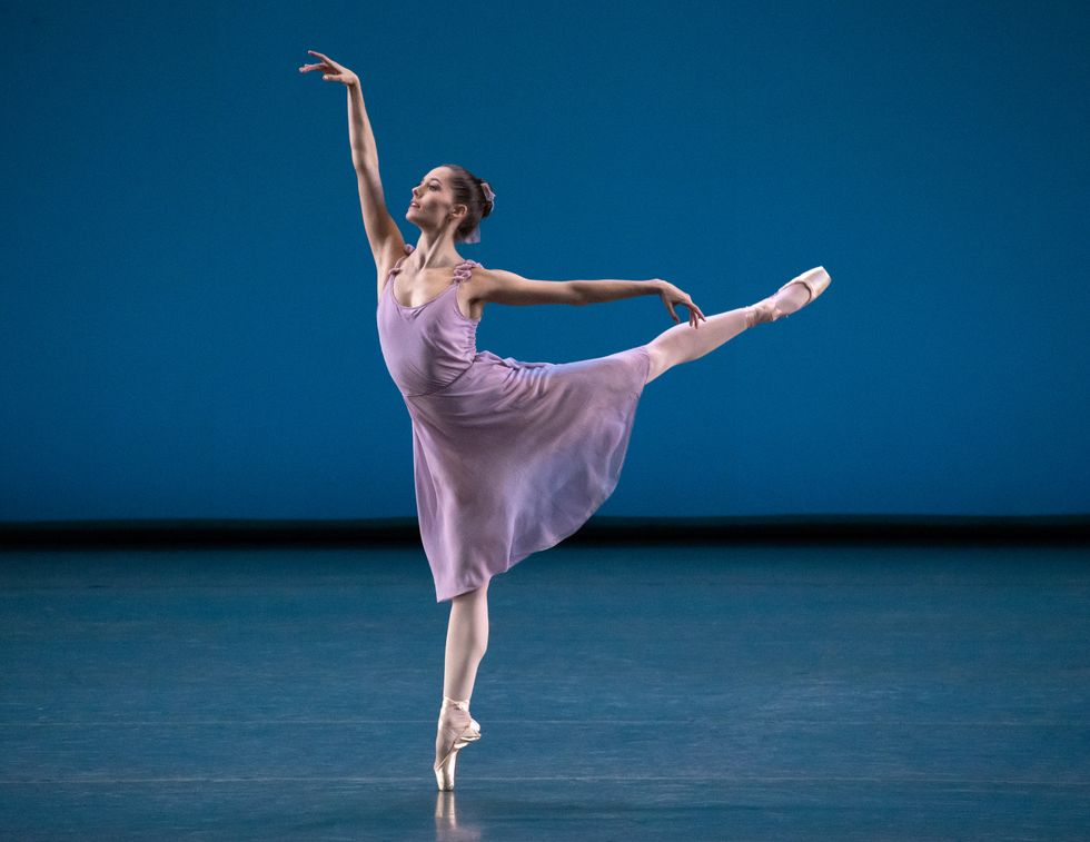 Phelan, in a light purple dress and bow in her bun, is in a luxurious first arabesque. She is alone onstage against a blue backdrop.