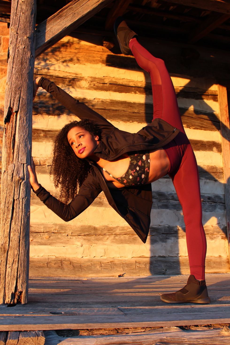 Moore in a penchu00e9, leaning against a support beam of a rustic, wooden building. She is wearing maroon leggings, a floral crop top and a black jacket.