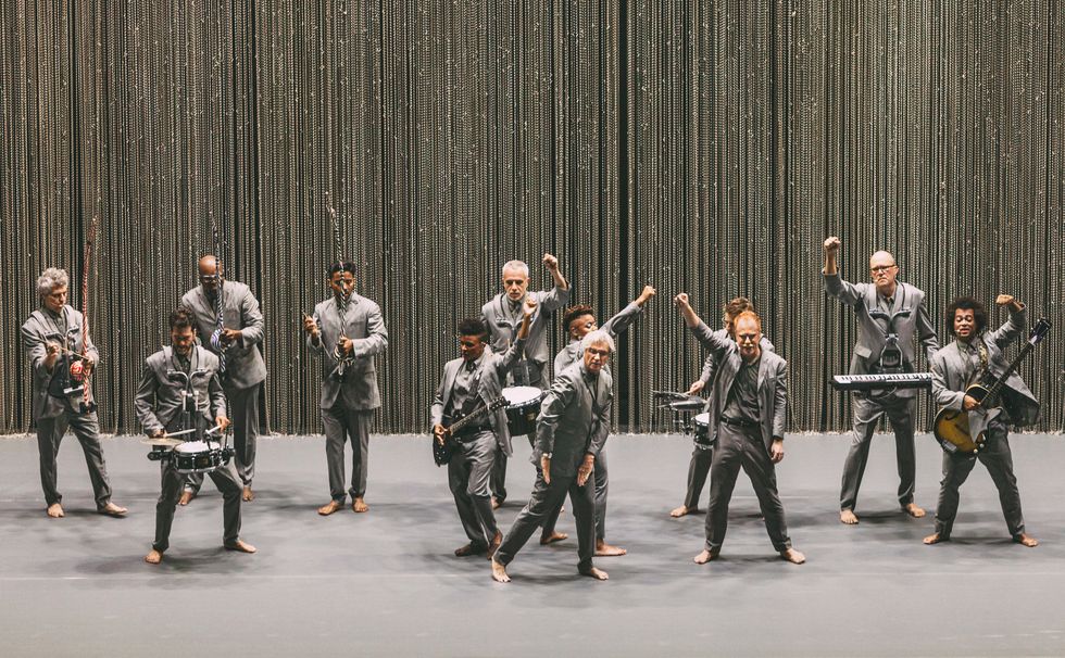 A group of 12 musicians wearing gray suits stand onstage. Most raise a fist in the air.
