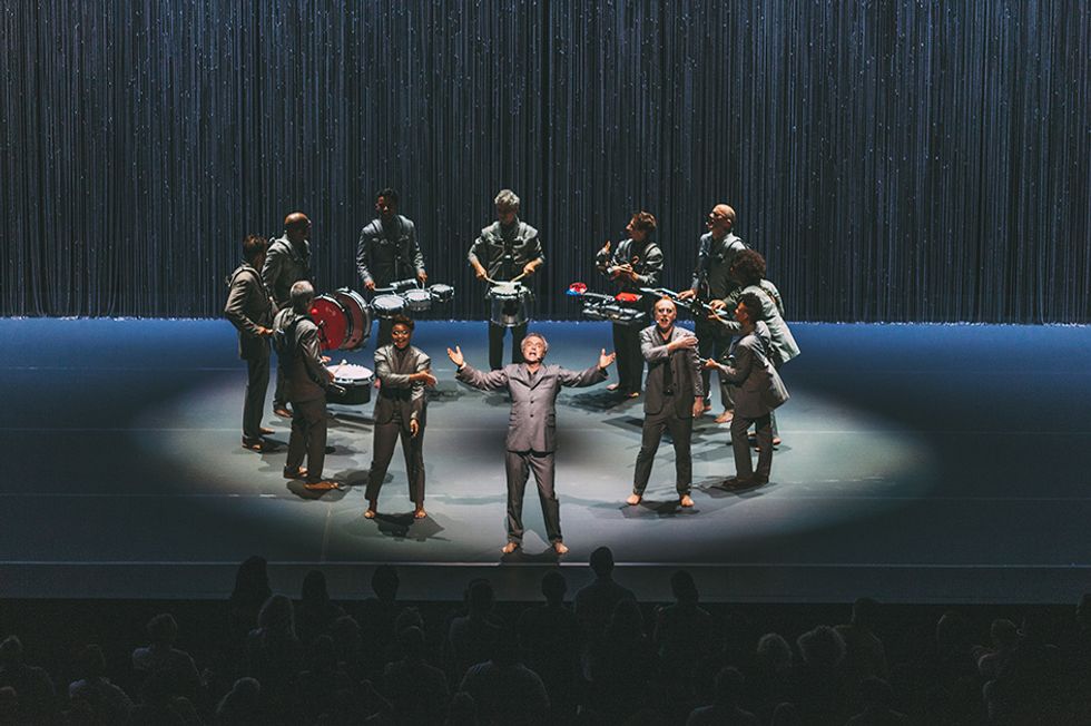 A circle of gray-suited musicians stands center stage. The man downstage center faces the audience and sings, his arms raised to shoulder height, palms open.