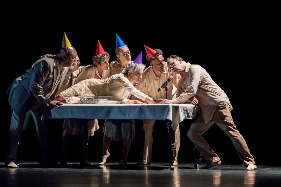 Six older performers in beige outfits and brightly colored party hats surround a table, one of them lying on one hip and reaching a hand toward a man whose head is inclined toward a microphone set at the edge of the table.