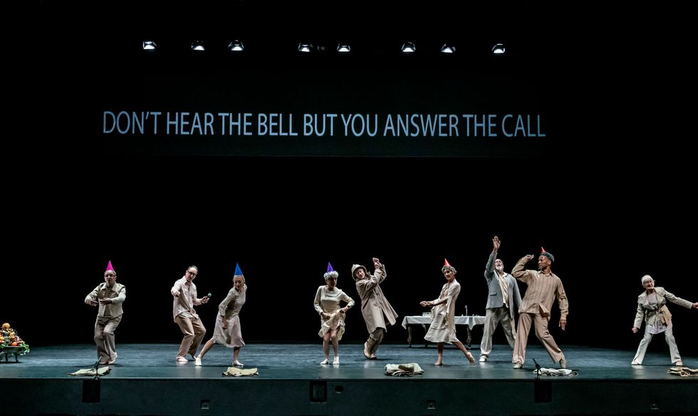 A group of nine older dancers are scattered around the stage. Half wear bright party hats, the others baggy coats. On the back scrim, the phrase "Don't hear the bell but you answer the call" appears in white in all caps.