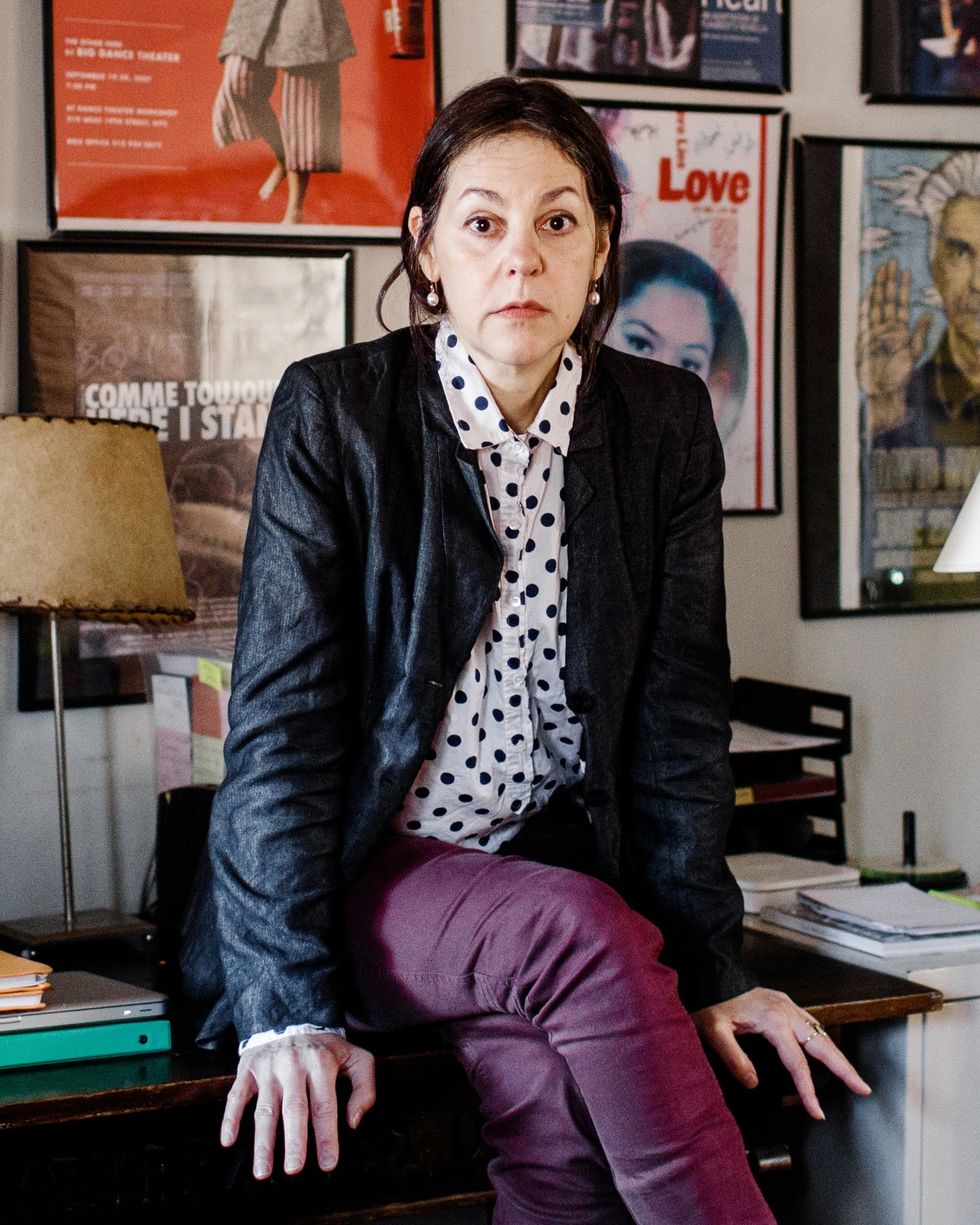 A white woman with dark hair sits on a wooden desk, looking questioningly at the camera. She wears a polka dot shirt, purple-red trousers, and a shiny black jacket. Framed posters line the wall behind her.