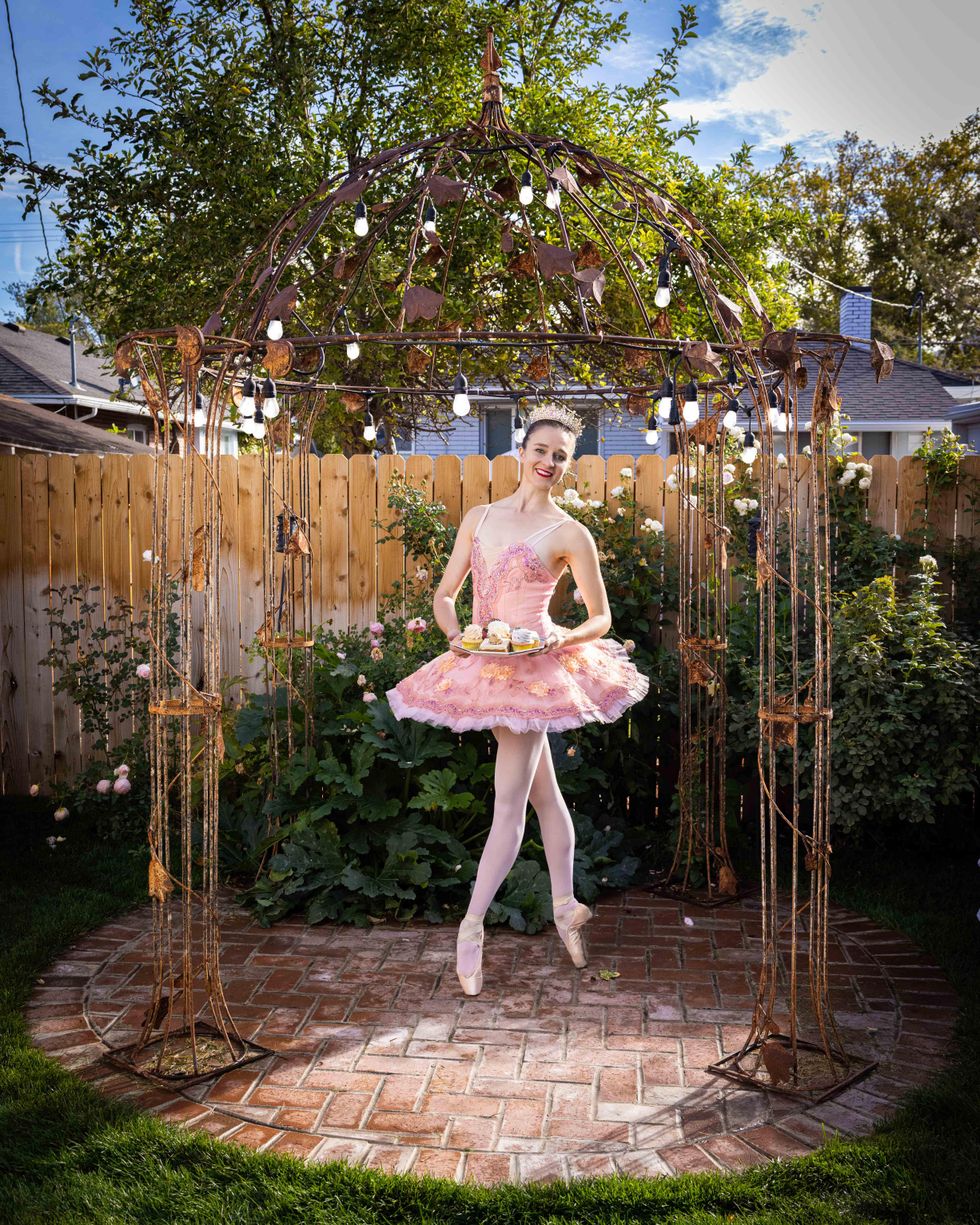 A white ballerina on pointe in fourth position in a tutu holds little cakes in a backyard