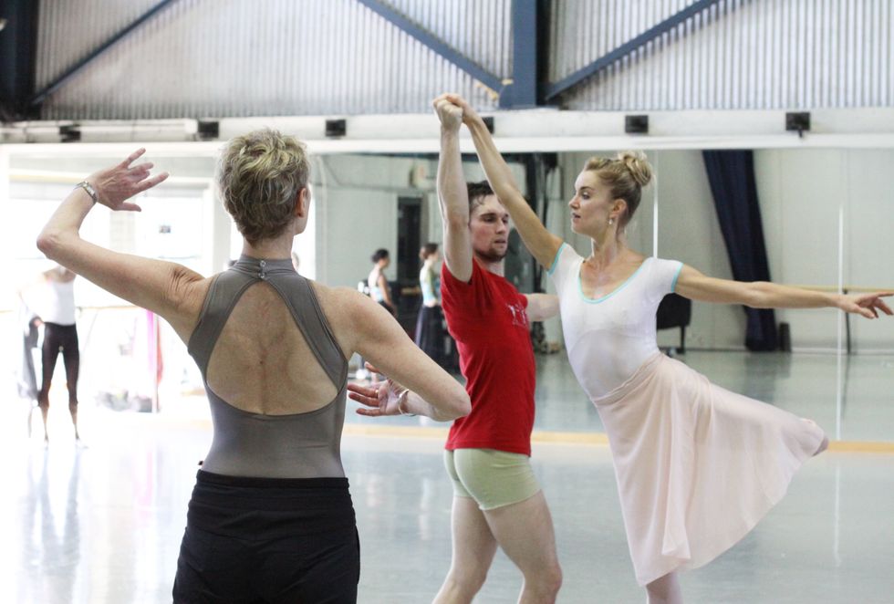 Kumery, seen from the back wearing a leotard and pants, gestures towards a couple rehearsing a pas de deux. The woman is in attitude back and the man partners her.