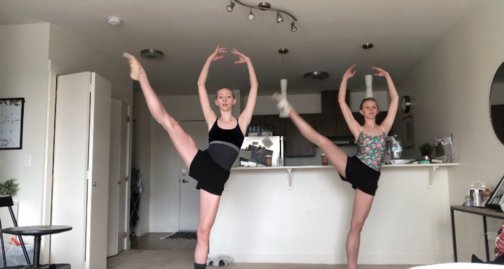 Two young female dancers in leotards and shorts practice du00e9veloppu00e9 a la seconde with their right legs, their arms in high fifth. They are practicing in front of their kitchen counter.