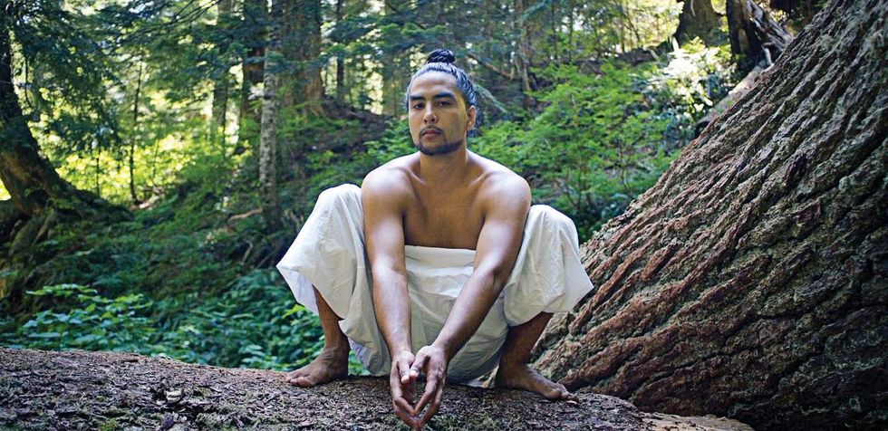 Dakota Camacho, barechested, crouches on a fallen tree in the forest, his hands touching in front of him as he stares at the camera