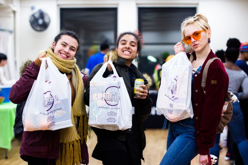 Three young women hold up bags of groceries, smiling.