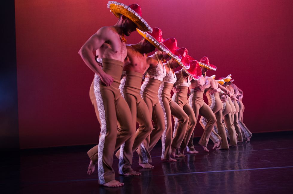 A line of male and female dancers in matching high-waisted brown trousers and bright pink and yellow sombreros stretches upstage. (The women wear lacy white halter tops.) The six closest to the camera face right, the rest left, all with arms linked around each other's waists as they step onto their right foot.