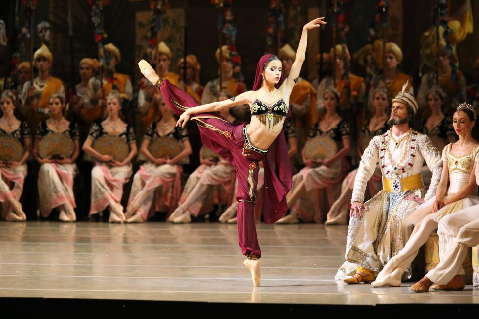 Khoreva poses en pointe with her right leg in arabesque. She wears dark red harem pants and a black brassiere with gold trim. Several lines of corps dancers stand and kneel behind her.