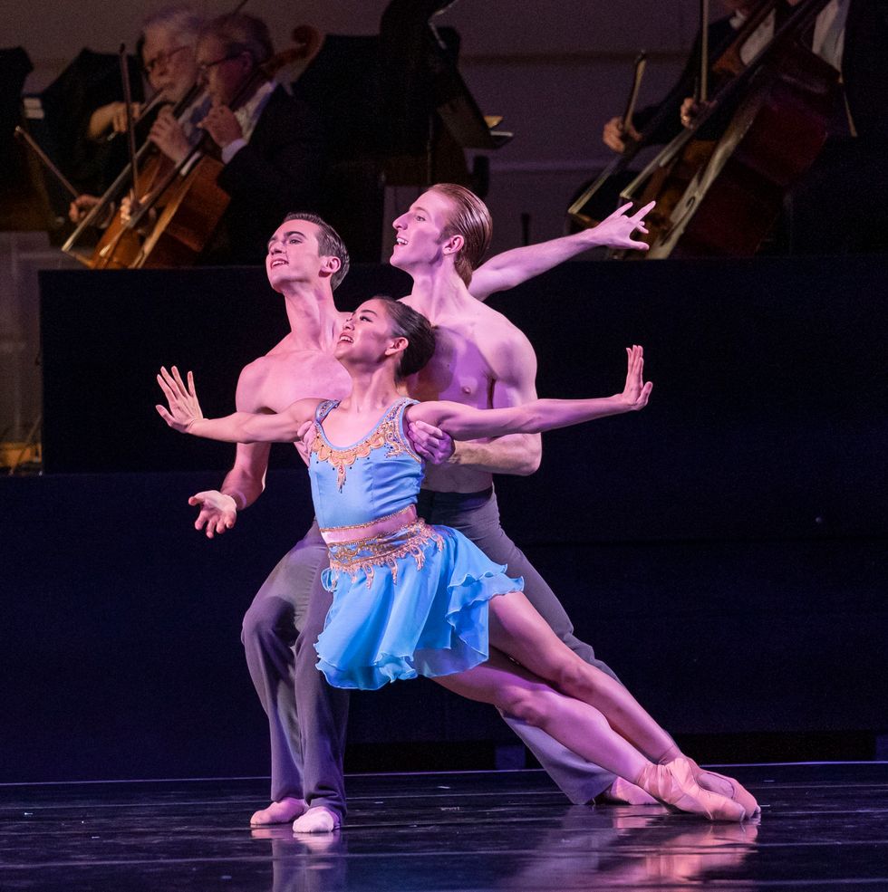 With her arms extended as she falls forward off pointe, Yumi Kanazawa, in a short, blue dress, is partnered by two shirtless men.