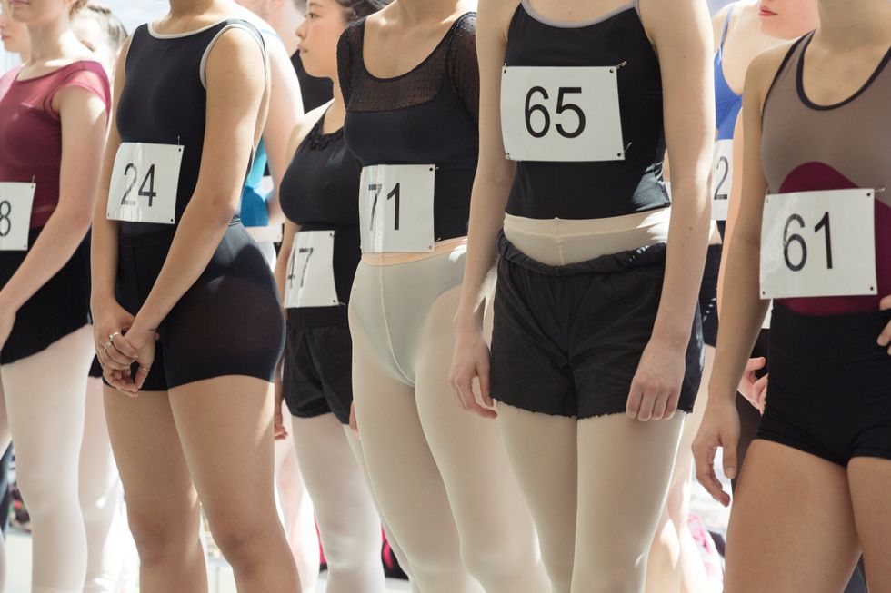 Auditioning dancers, wearing leotards and audition numbers pinned on to their clothing, are photographed from the shoulders down.