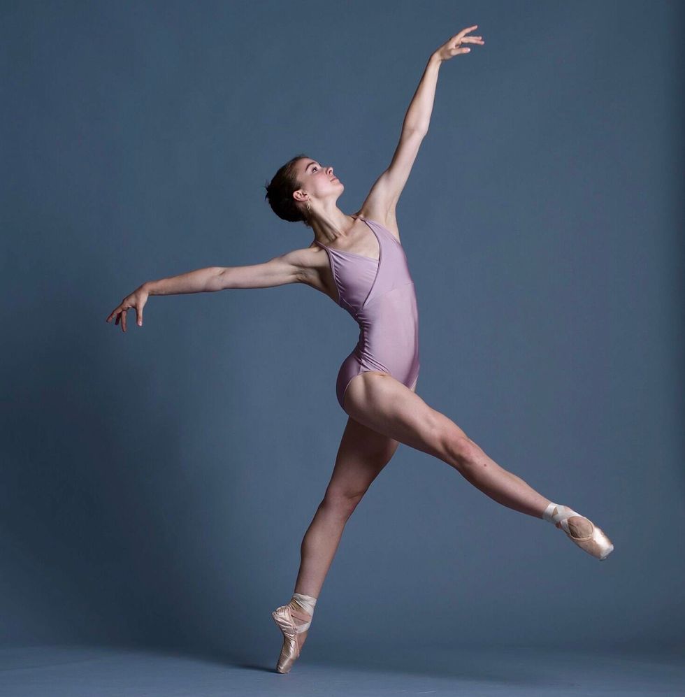 Peabody posed on pointe with one leg extended forwards and her arms reaching long. She is wearing a lavender leotard and the background is gray.
