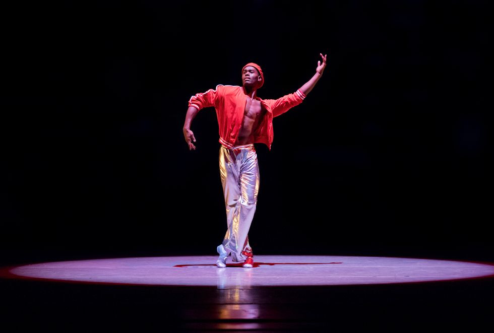 Roberts stands on a dark stage, lit by a spotlight. He is wearing silver pants, an open red bomber jacket and a red hat. His arms are curving in opposite angles.