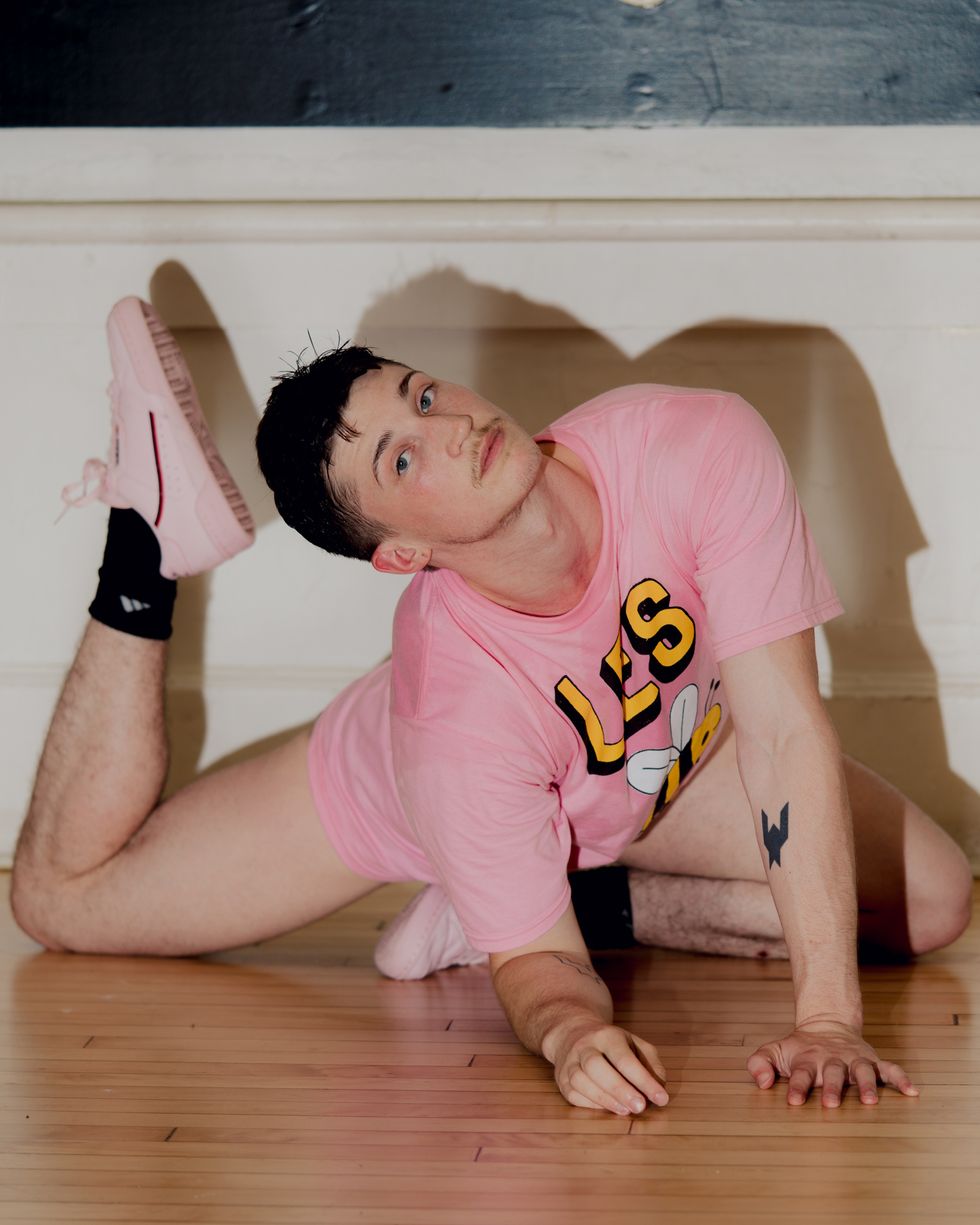 A young, white man poses on a wooden floor. He kneels on one shin, while his other leg is extended behind him and bent at the knee, his lower thigh resting on the floor. He leans forward onto one hand and one forearm as his head tips toward his back foot. The oversized pale pink graphic t-shirt he wears matches his sneakers.