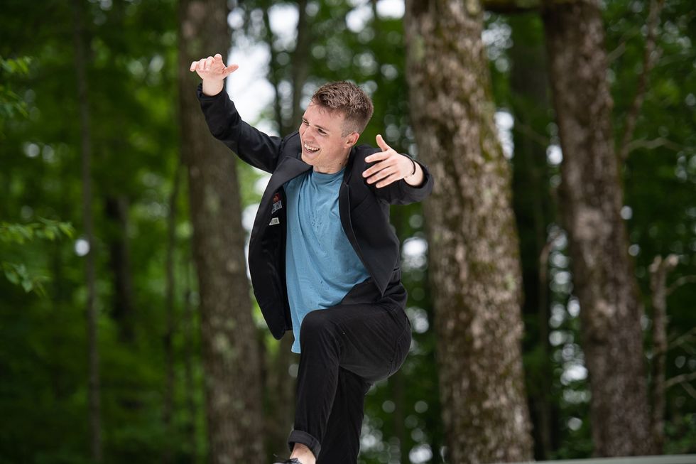 A young white man wearing a suit jacket over a blue T-shirt grins as he balances on one leg, arms flung up around his head in an embracing gesture. Trees and greenery are blurry but visible in the background.