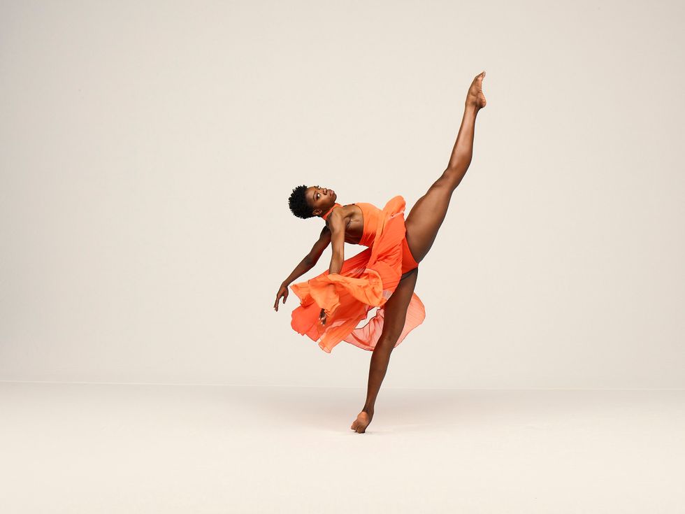 A long-legged, dark skinned dancer, looks calmly at the viewer as she balances on one leg. She is situated in profile, her legs extended to a 180-degree layout on relevu00e9, her torso nearly parallel to the floor, as her orange dress flairs behind and below her.