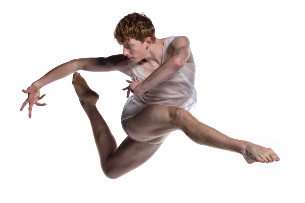A red-haired man leaps, legs bending to parallel attitude in front and back as he twists around to his back leg, arms curving at shoulder level.