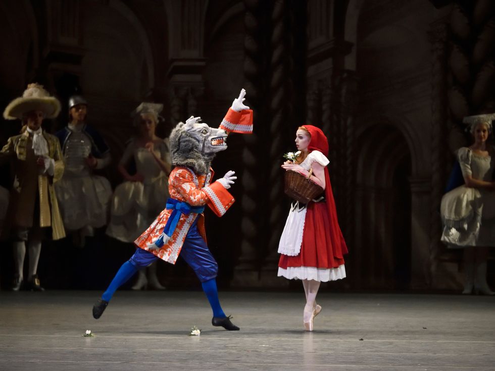 A young woman in pointe shoes, dressed as Little Red Riding Hood, looks startled as she hovers on pointe and someone in a wolf costume gestures toward her. Courtiers look on in the background.