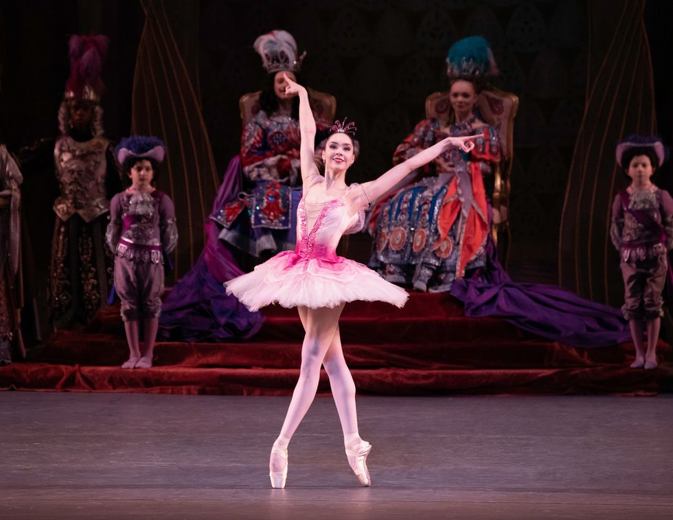 A statuesque dancer in a classical tutu and pink tights balances in fourth position croisu00e9 en pointe, arms extended in an asymmetrical "V" with index fingers extended.