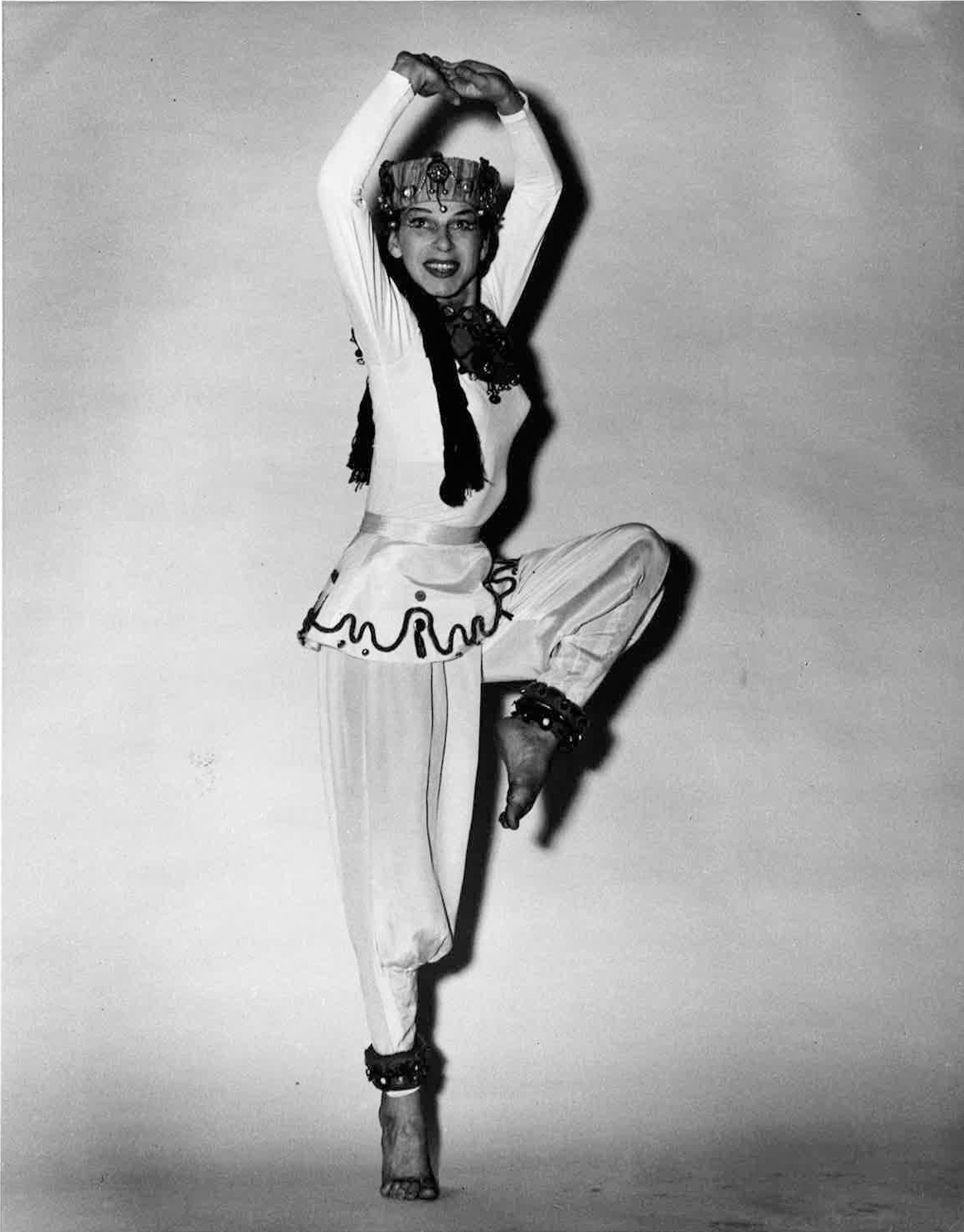 A barefoot Anna Halprin grins past the camera as she balances on relevu00e9 on one foot, the other leg raised to 90 degrees and bent at the knee and slightly turned out as her arms stretch overhead, fingers clasped.