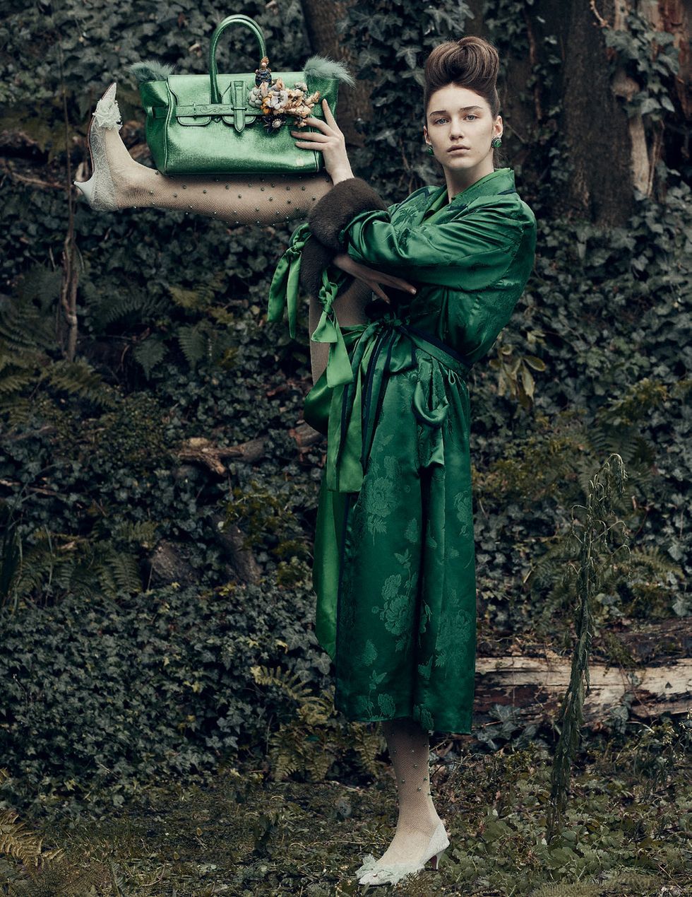 A model in a green dress holds up a purse with her shin, her leg lifted up to her ear