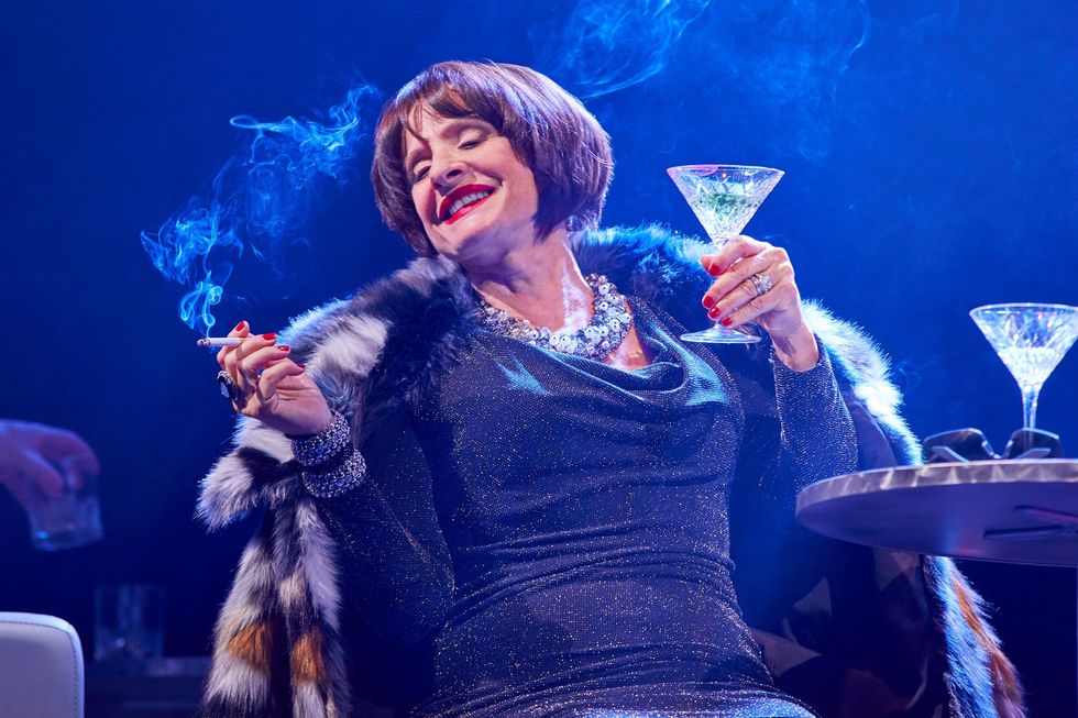 Patti LuPone in costume as Joanne, in a fur coat, large necklace and bracelets and sparkly black dress. She is holding a martini glass in one hand, a cigarette in the other, as she smiles.