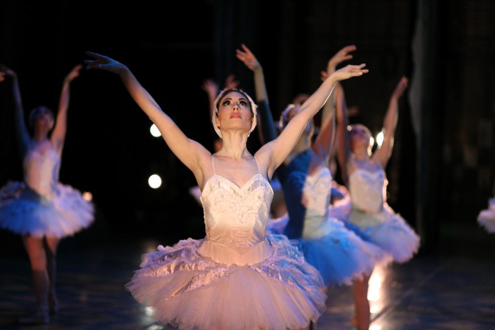 Morgan Butler extends her arms, swan-like, on either side of her head, looking up above her. Other dancers as swans are blurry in the background
