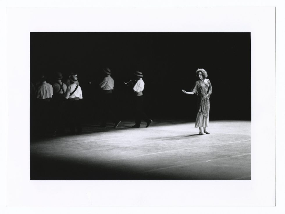 In a black and white archival photo, Trisha Brown stands on a bare stage, looking to her right hand as it extends to the side, elbow bent and palm up. In the background, a group of men in boater hats and suit pieces walk away.