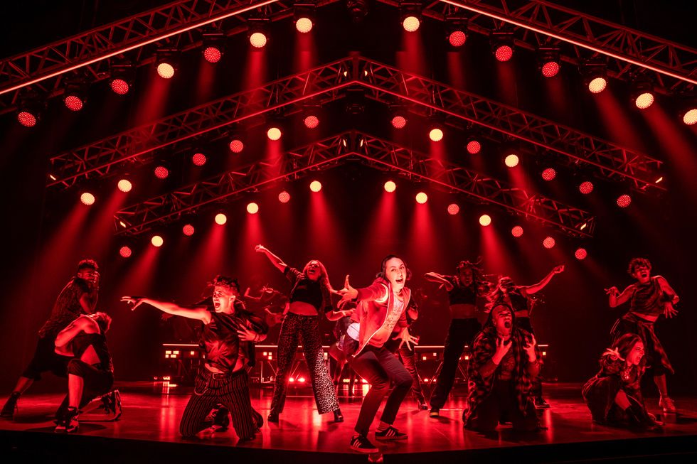 On a red-lit stage, a dozen performers wearing casual clothes punch the air and scream while standing or kneeling. Lauren Patten crouches at center just before the orchestra pit, reaching for the audience as she sings.