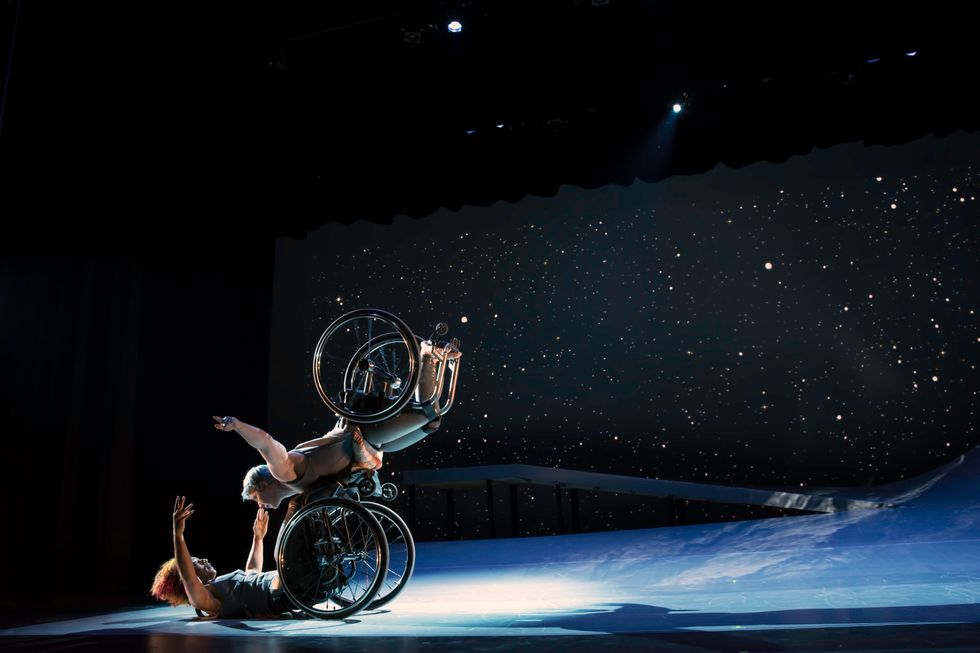 Laurel Lawson as Venus flies in the air with arms spread wide, wheels spinning, and supported by Alice Sheppard as Andromeda who lifts from the ground below. They make eye contact and smile. A starry sky fills the background and moonlight glints off their rims.
