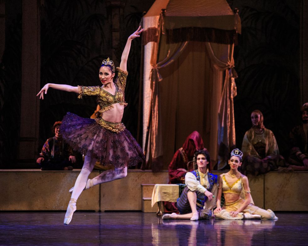 In the foreground, a female dancer in an opulent tutu and cropped blouse is caught mid air in a petit jete. In the background, a male and female dancer recline on the floor together and watch.