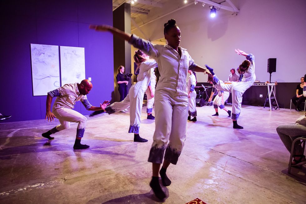A group of dancers performing in a museum-like setting. They wear what look like white painter jumpsuits, with blue patterns on the bottom of the pants and the arms. They are in various positions, with the audience sitting and standing around.