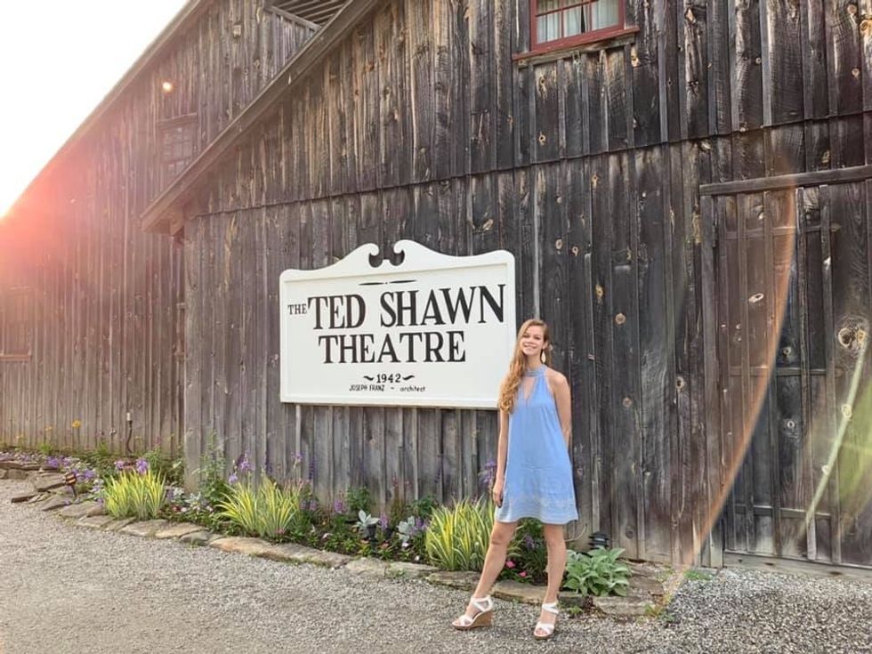 Madalyn Rupprecht smiles as she stands in front of what looks like a barn made of dark, worn wood, bearing a large white sign reading "The Ted Shawn Theatre."