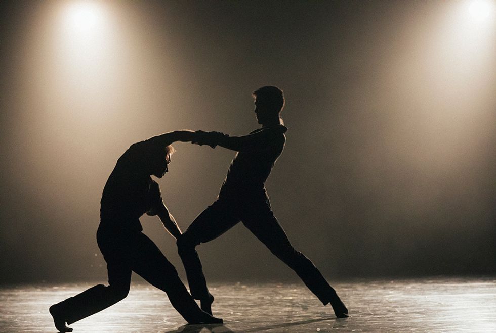 Two male dancers are seen in silhouette on a hazy stage. Their hands are clasped as they lunge toward each other, one dancer slumping forward to extend his free arm to the other's bent knee.