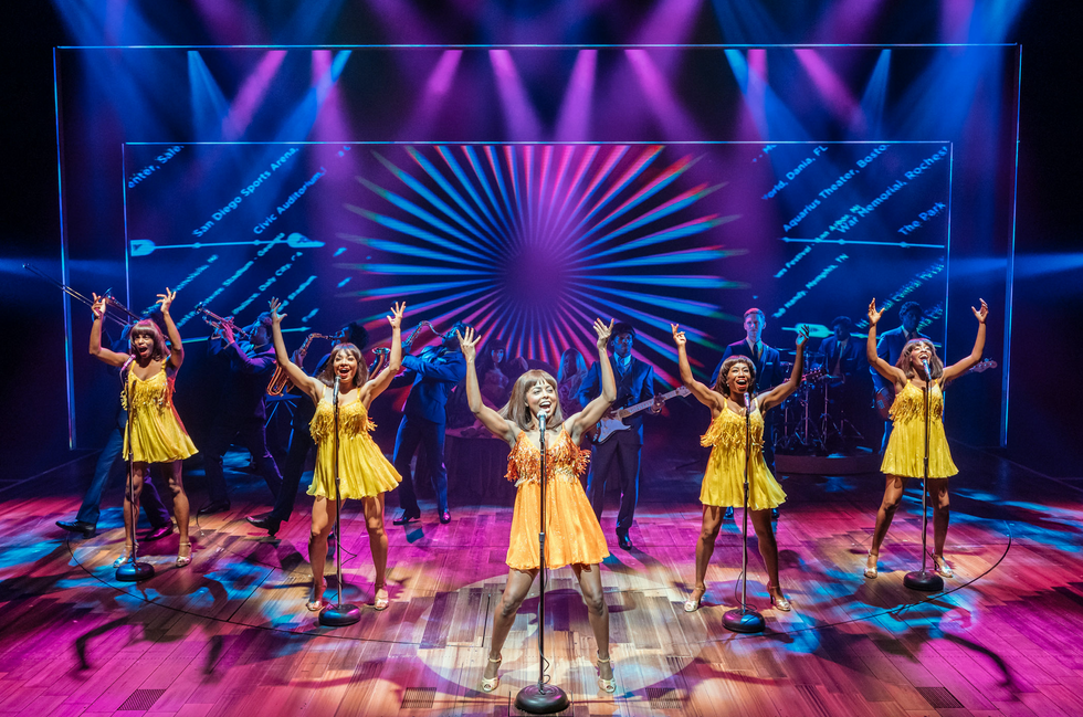 Five women in yellow dresses stand in front of microphones on a brightly lit stage, their hands in the air as they sing. A band is behind them.