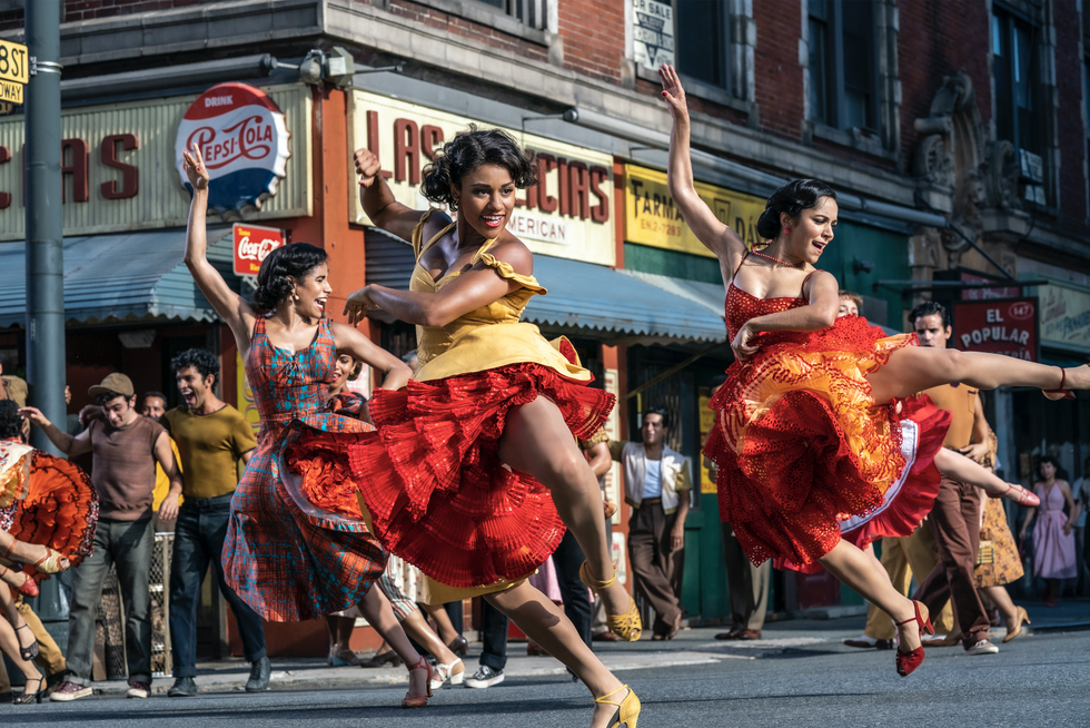 Ariana DeBose wears a yellow dress with red ruffles underneath, kicking her leg to the side. Two dancers behind her mirror her movements.