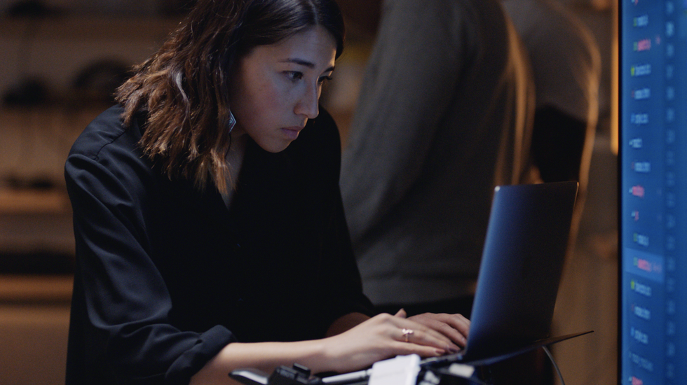A young woman with her shirtsleeves cuffed to the elbow looks intently at a laptop, fingers at the keys.