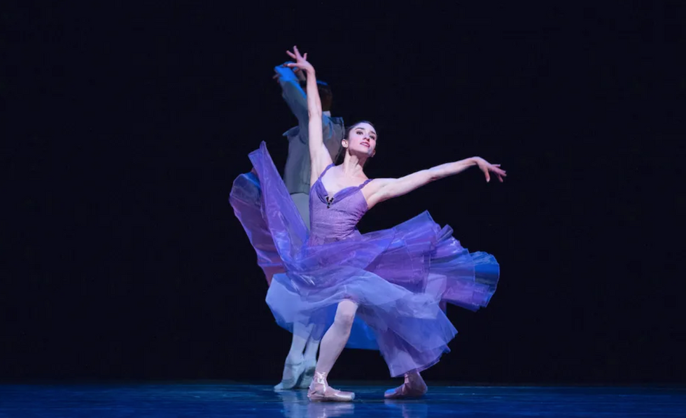 Biasucci onstage in a long, flowy light purple dress and pointe shoes. She is in a wide fourth position, with one arm above her and one arm to the side, and an exaggerated head. The background is black.