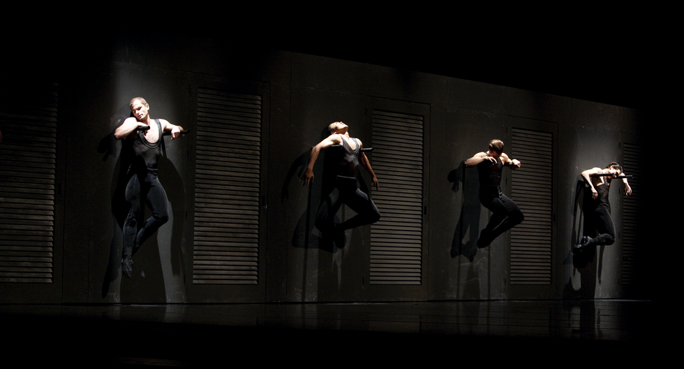 Four male dancers hang from a wall, all in black