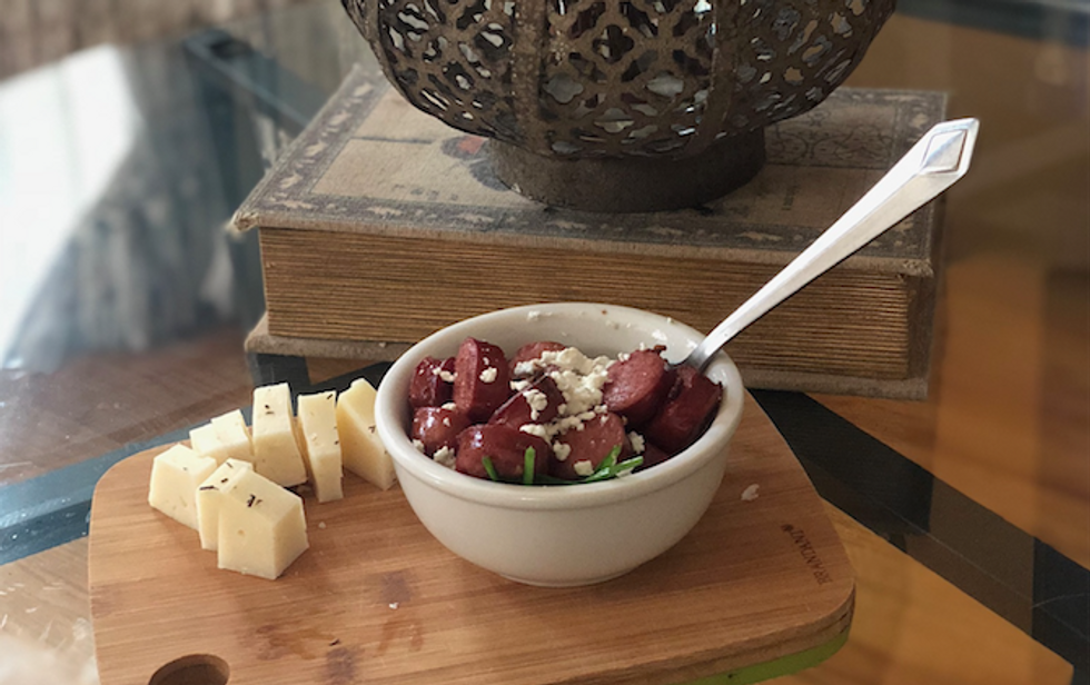 A bowl of kielbasa with veggies and goat cheese, sitting atop a cutting board with pieces of cheese