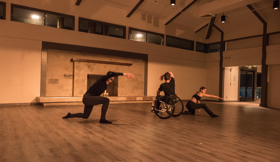 Two dancers gather their outstretched arms over their head as they pull forward from the ground into a deep lunge. Between them, a third dancer in a wheelchair does the same while facing toward the back. The trio wears a variety of black tops, dark grey pants, and black socks. They are dancing on a slippery, brown laminate floor in a large community hall with tall, white ceilings and a large, taupe fireplace behind them.