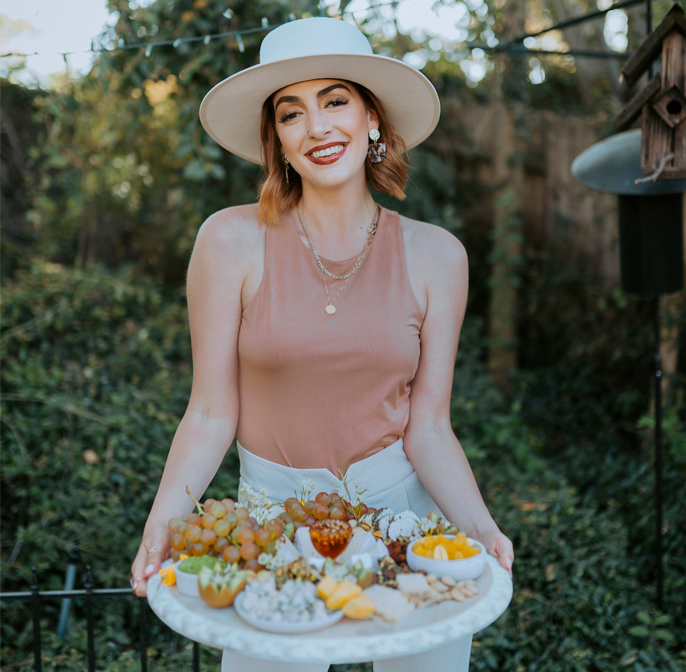 Chelsea Farrah, in a big white hat, holds a platter of food in front of greenery