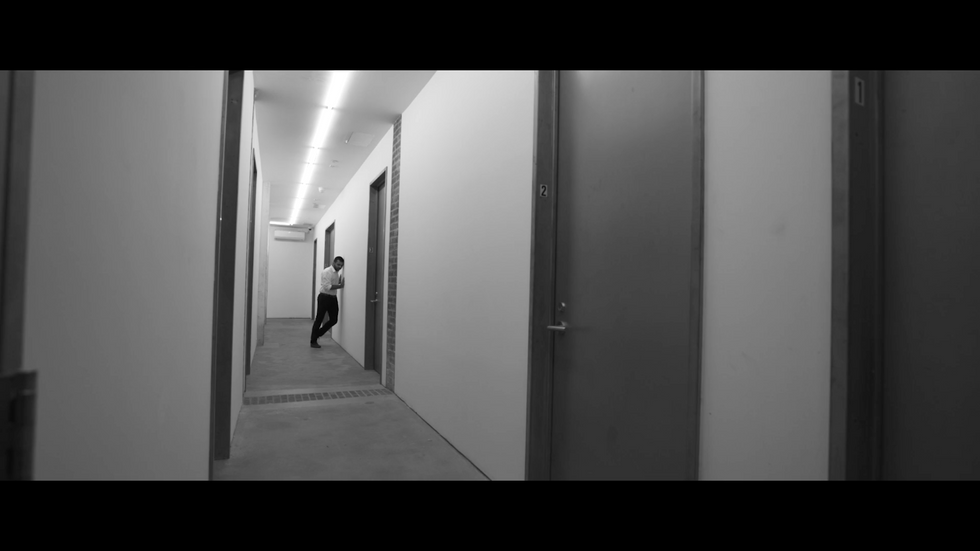 Black and white wide-screen shot of a long white hallway with grey doors, one man leaning against the wall in the distance