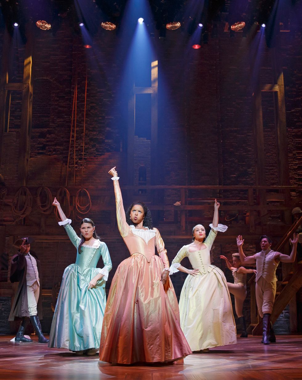 Phillipa Soo, Renu00e9e Elise Goldsberry and Jasmine Cephas Jones each extend one arm overhead as they snap, leaning into their hips. They wear shiny, pastel-colored dresses that nod to Hamilton's late 18th-century setting.