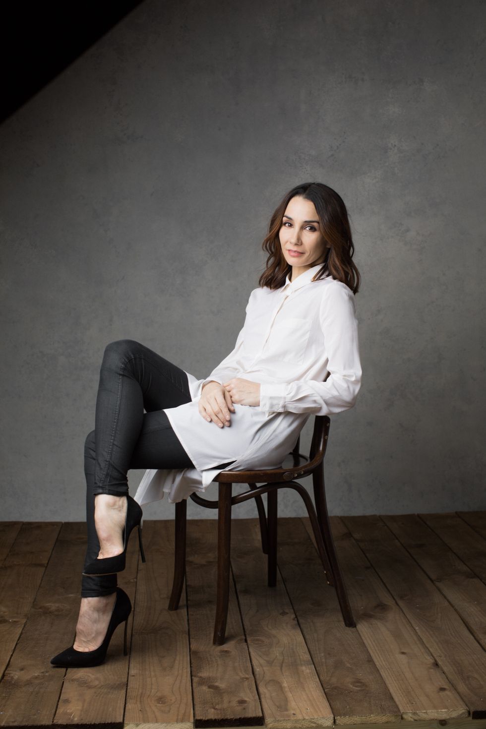 Tamara Rojo, brown hair in loose curls around her face and wearing a white button down, black trousers and high heels, reclines elegantly in a wooden chair.