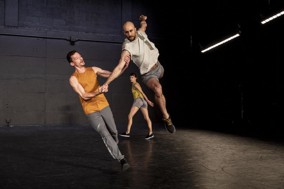 In the foreground, two male dancers are photographed in middair while leaping forward and using each other's arms for support. In the background, a female dancer looks on midstep.